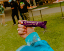 Load image into Gallery viewer, Hand holding a lavender refreshment towel package in an outdoor setting with people and tables in the background.
